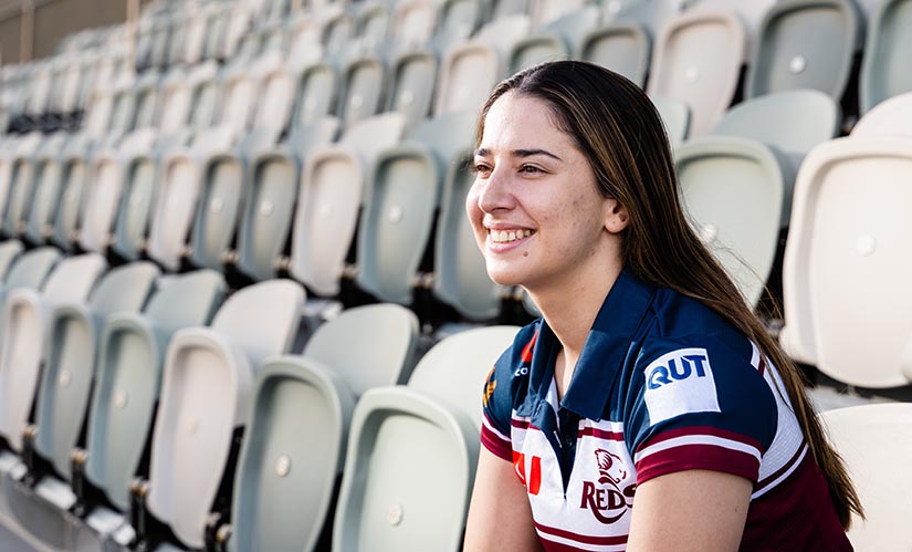 A young woman wearing a Queensland Reds polo shirt sits in a stadium.
