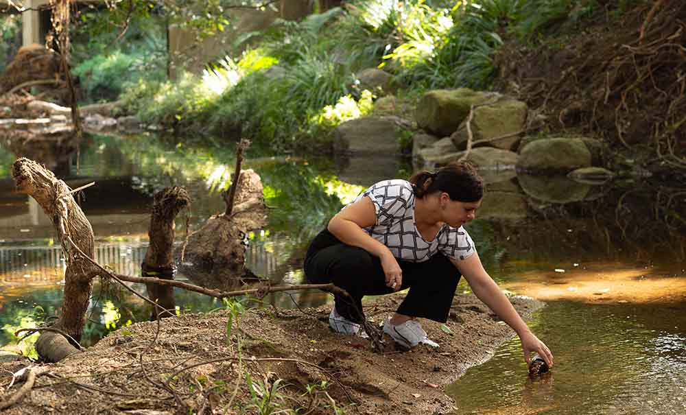 A woman crouches beside a shallow creek, she is dipping a small jar into the water to collect a water sample.