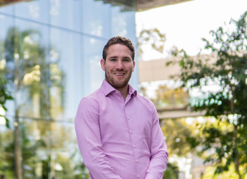 A smiling man in office attire stands in front of a glass building facade.