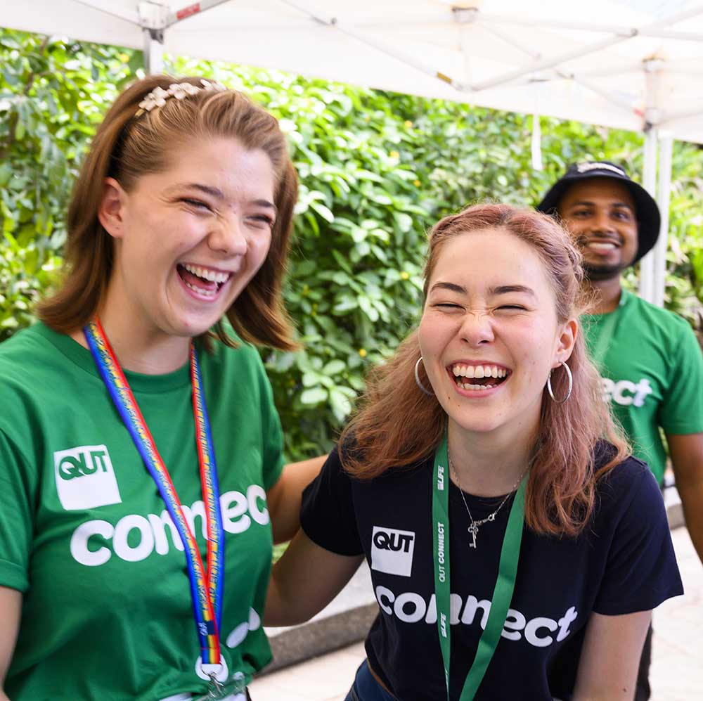 Two female students wearing QUT Connect t-shirts are laughing while working at an outdoor event, while another student looks on.