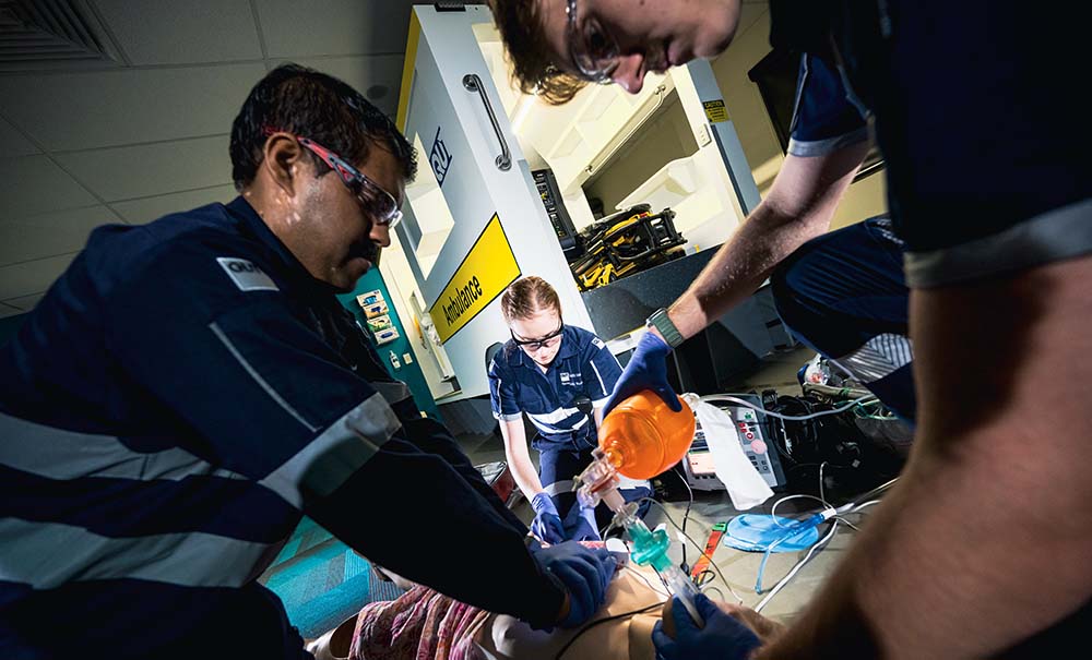 Three students, including Megan, work to revive a dummy during a training scenario. They are crouched on the ground around the dummy, they are using equipment and vials of saline that simulate medicine.