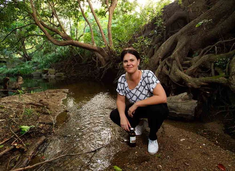 A woman holding a sample bottle crouches on the bank of a shallow creek.