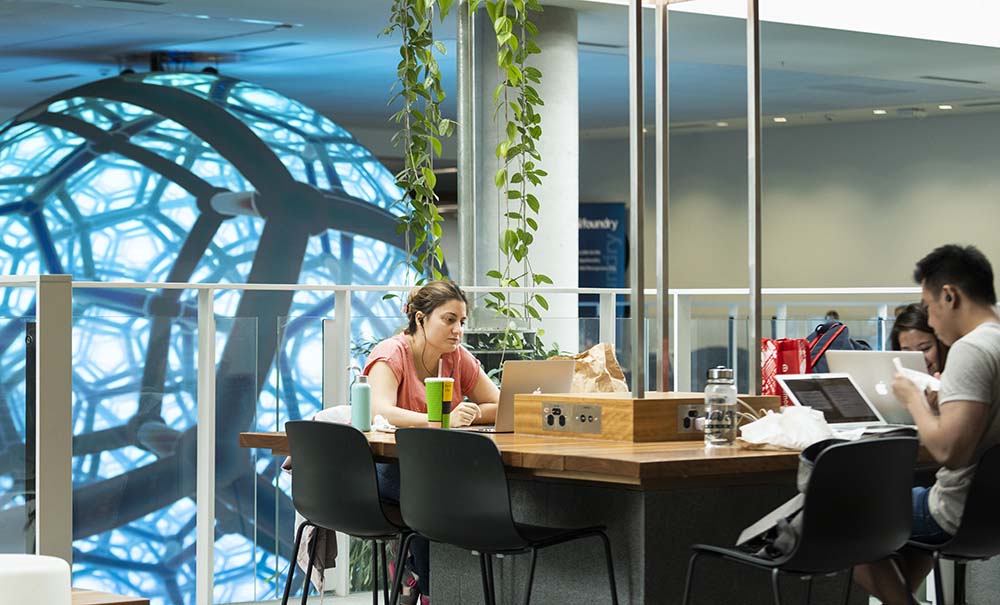 A woman sits at a table in a study area on campus, in front of her is a laptop and she is listening to a video through her headphones.