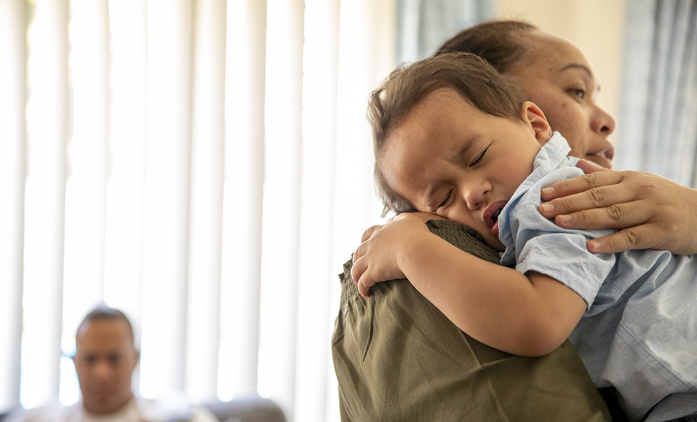 A mother holds and comforts a crying toddler while dad sits in the background.