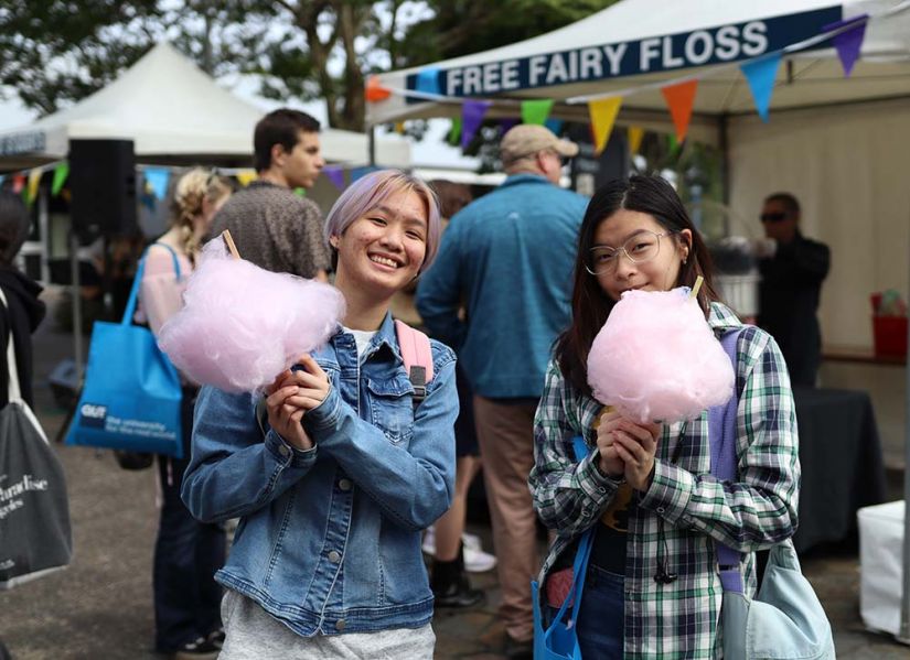 Two smiling students holding fairy floss stand in front of a brightly coloured tent.