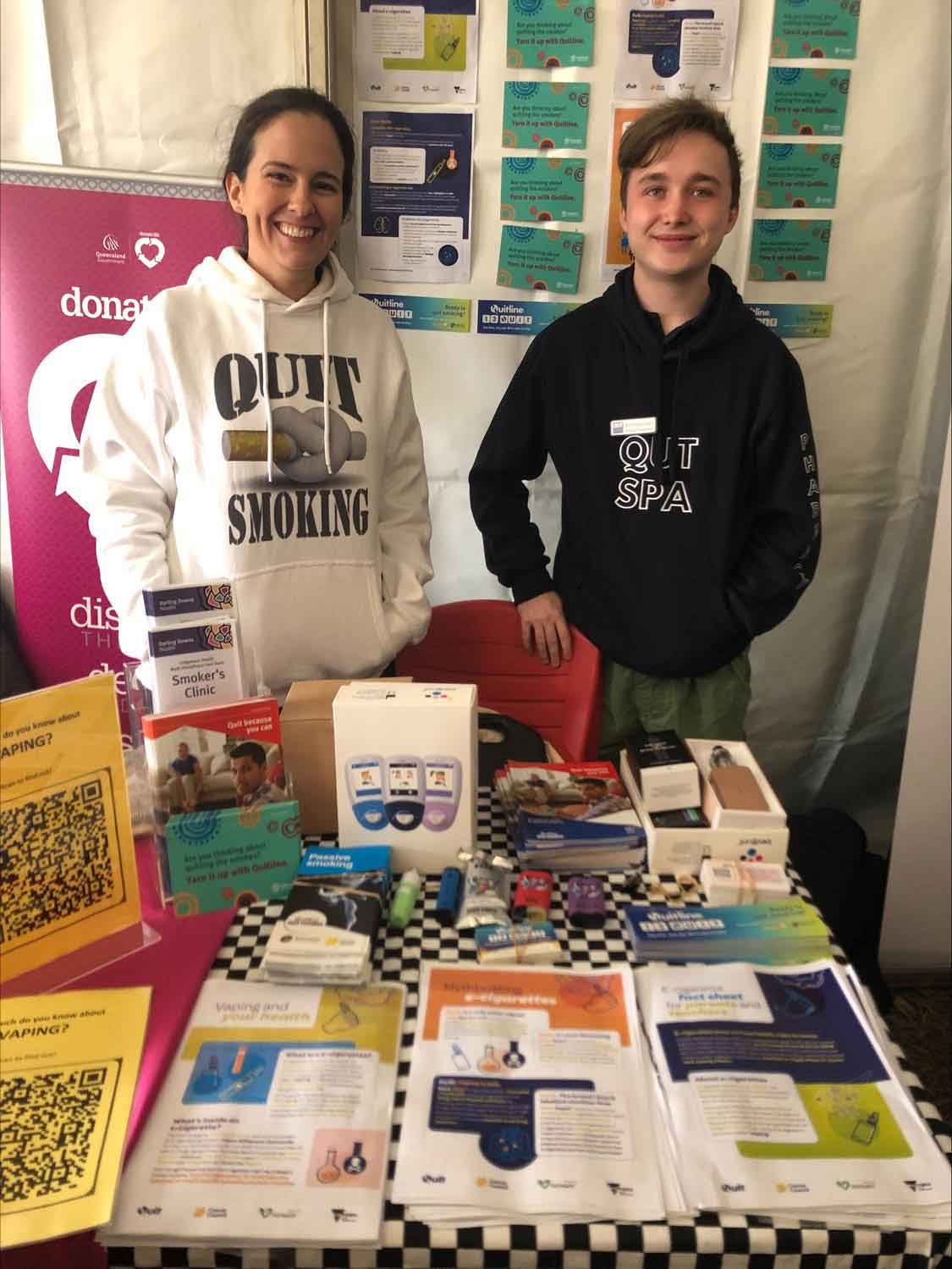 Two phamacy students a male and a female wearing 'Quit smoking' shirts stand behind a table displaying pharmacy products at an industry event. 