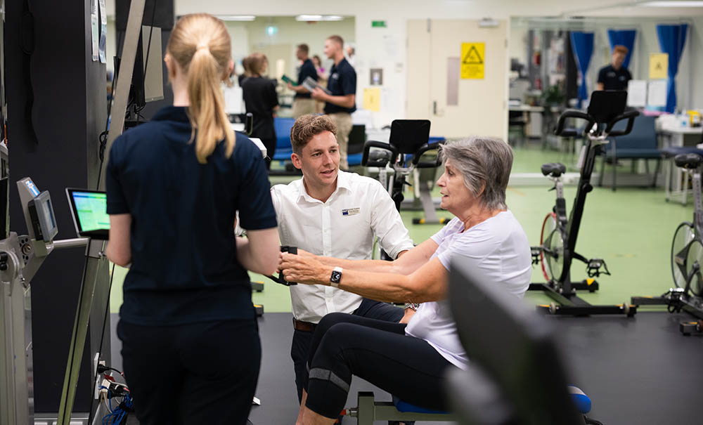 A smiling teacher supports a student and a client as they move through prescribed exercises.
