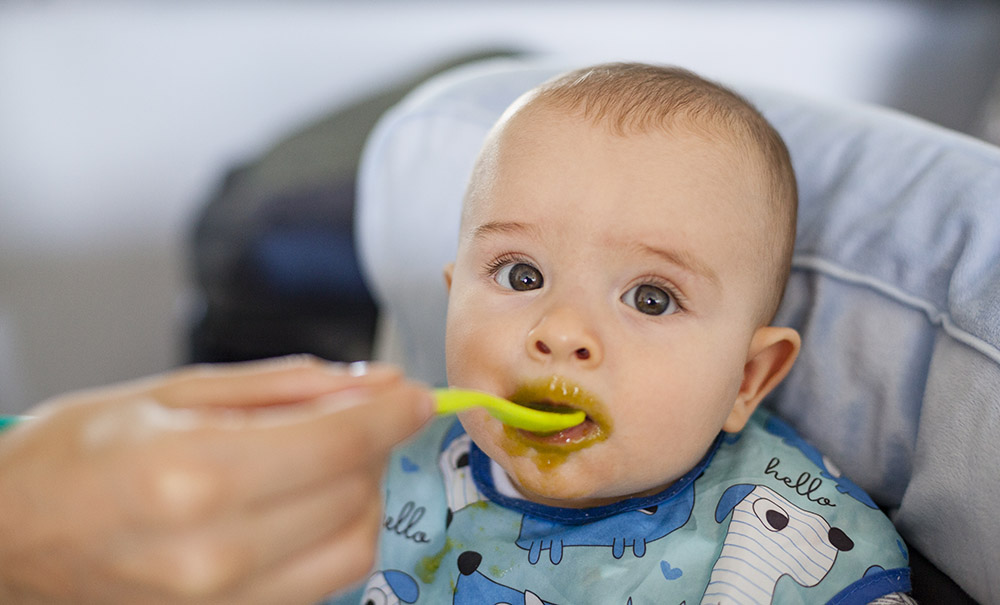 A messy-faced baby is spoon fed green baby food.