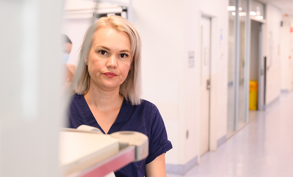 A frowning nurse with shoulder length hair stands in a hospital hallway and stares into the distance.