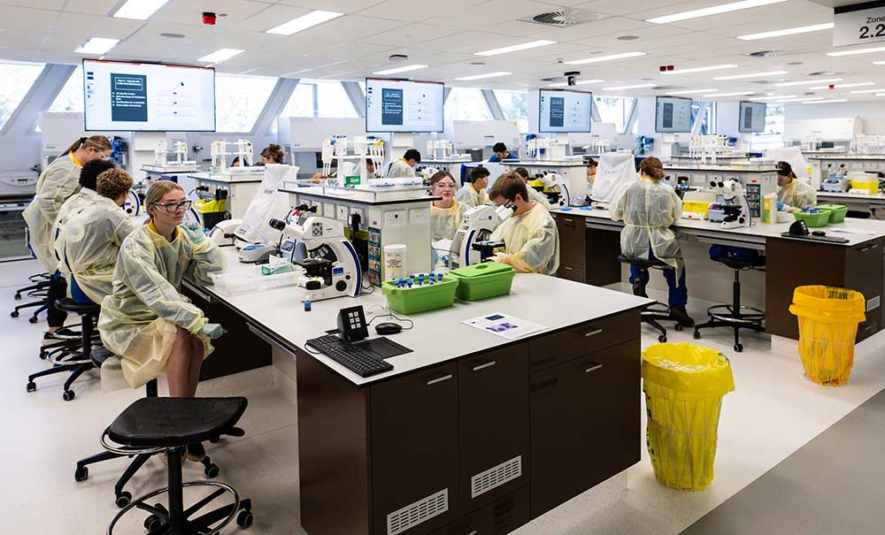 A class of students wearing lab coats, gloves and glasses sit at rows of benches in a modern, brightly lit laboratory.