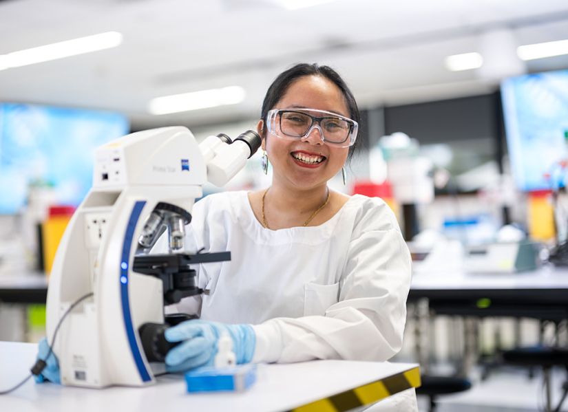 A female student wearing a labcoat and protective glasses sits infront of a mircroscope in a modern laboratory.