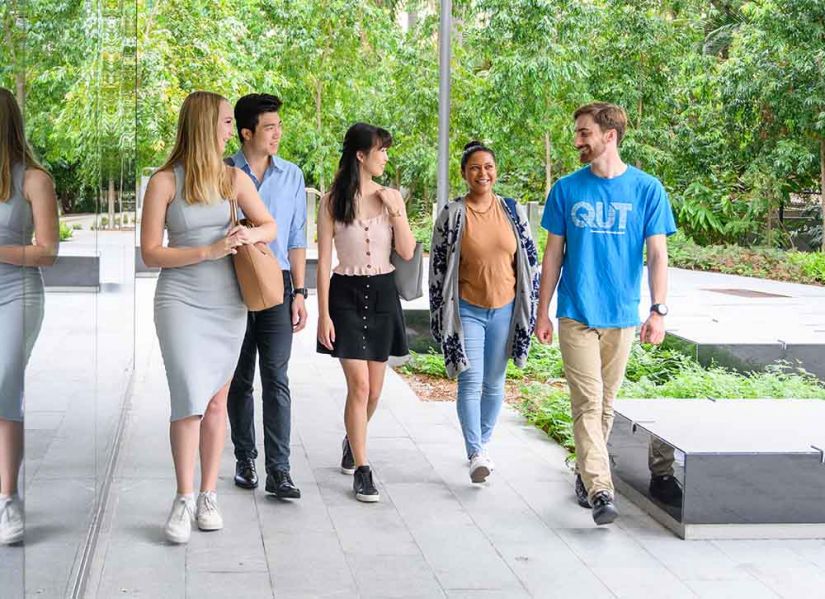 A group of students from a range of backgrounds listen to a QUT guide as they tour the campus on a sunny day.