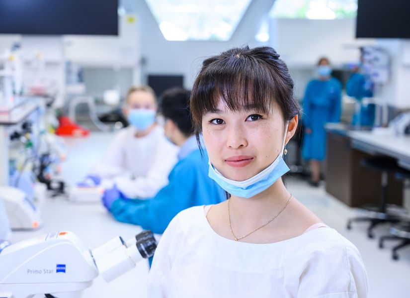 A woman wearing masks and a lab coats sits at a laboratory workbench.