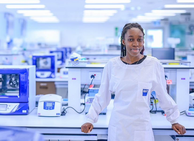 A female student with her long braids pulled back, wearing a QUT lab coat, standing at a bench in a large, well equipped laboratory. 