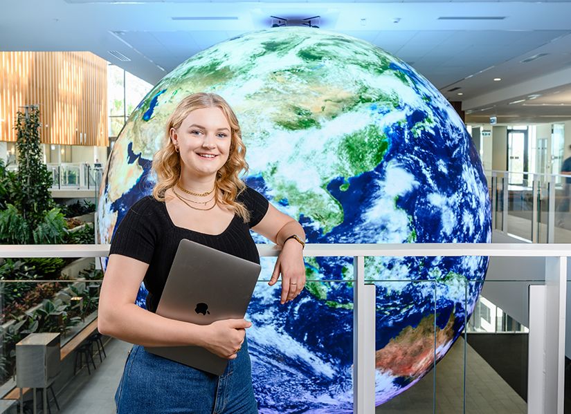 Young woman posing in front of a large digital sphere displaying a realistic artwork of the Earth. She is holding a laptop and smiling towards the camera.