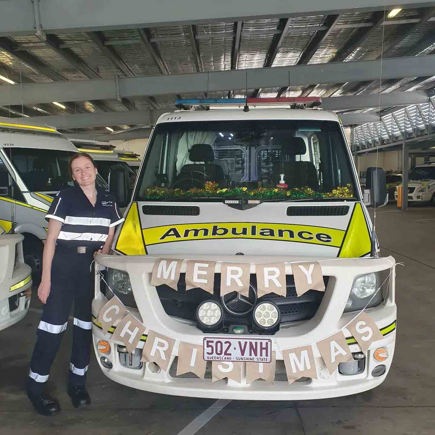 Megan wears a paramedic uniform and stands next to the front of an ambulance decorated with banners that read 'Merry Christmas'.