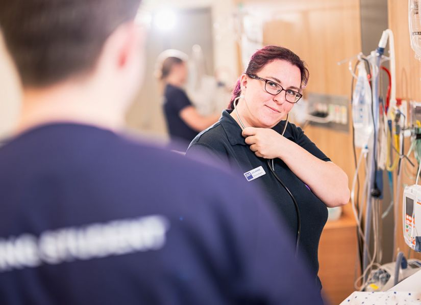 A female nursing student looks up and pulls a stethoscope out of her ear to hear what is being said to her by another student.