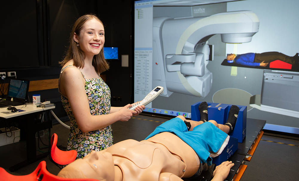 A smiling student stands next to a gurney with a mannequin patient. Behind them is a giant screen with a simulation of radiation therapy treatment.