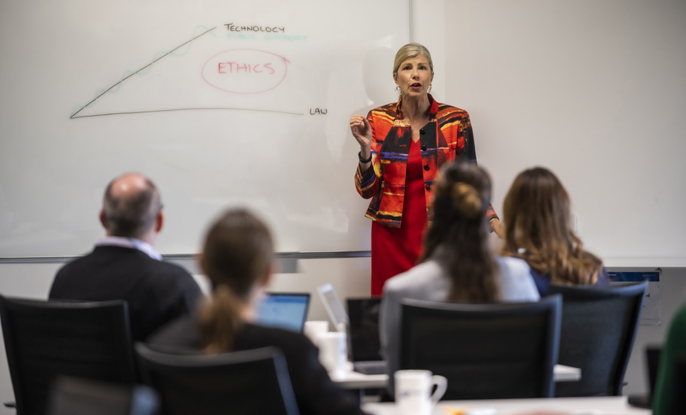 A woman stands in front of a white board talking to a small group of professionals seated at tables with laptops.