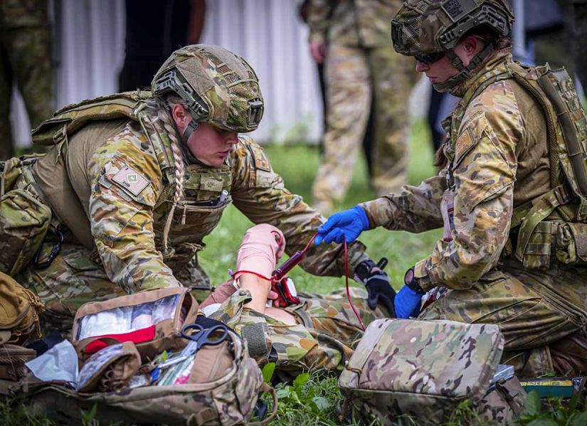 Two army paramedics in full camoflage and kit crouch on the grass to treat a patient in a practice scenario.