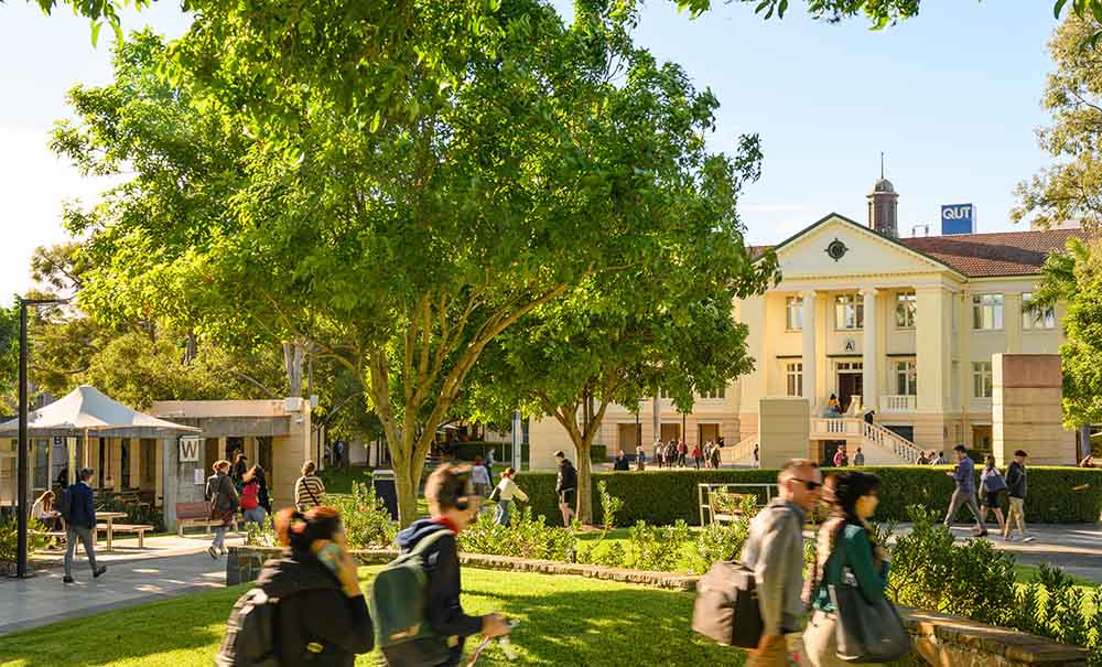 A group of students walk past trees on an open, leafy university campus. In the background is an old stone Queensland school building.
