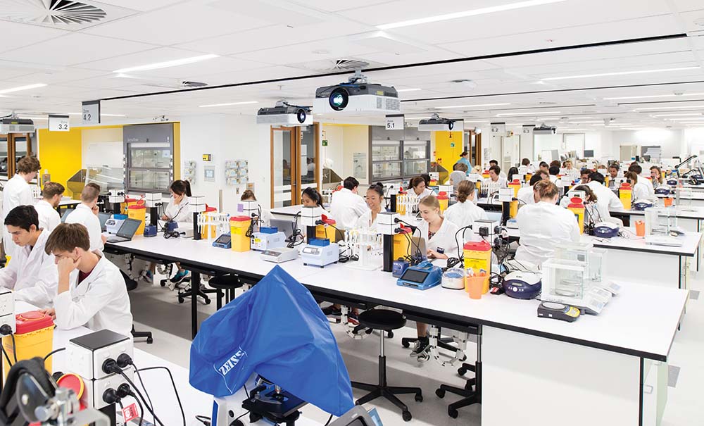 In a large laboratory space many students wearing protective clothing sit at rows of benches.