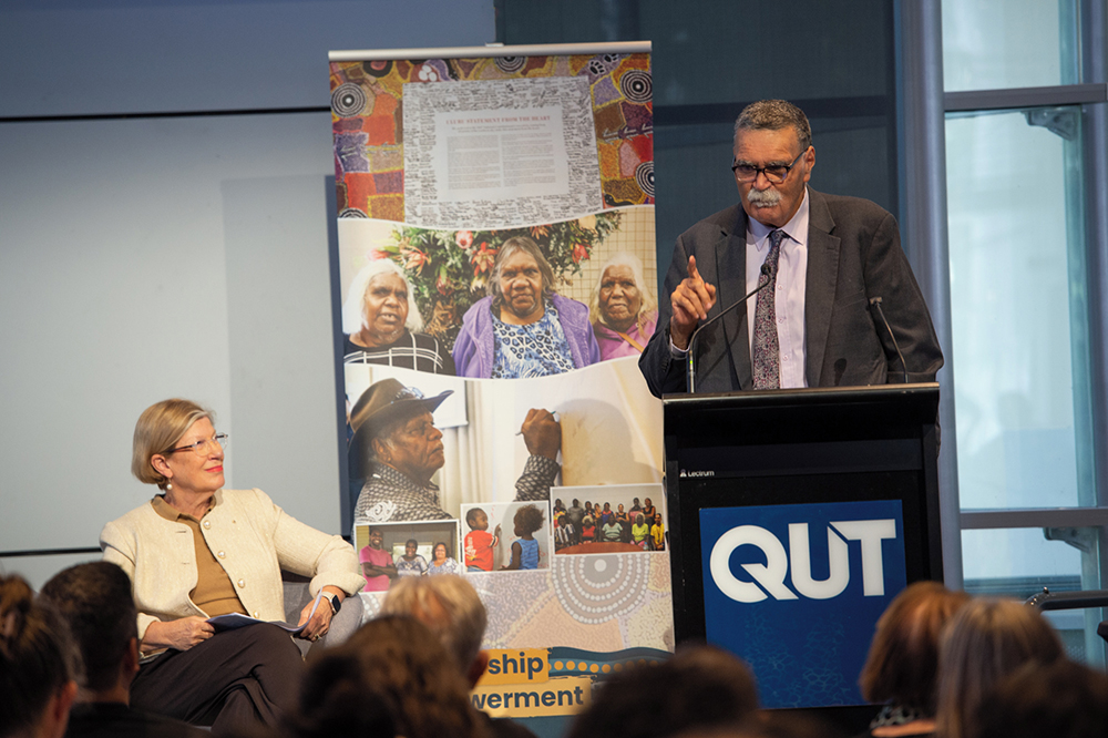 White woman looks towards male Aboriginal elder speaking at a QUT lectern