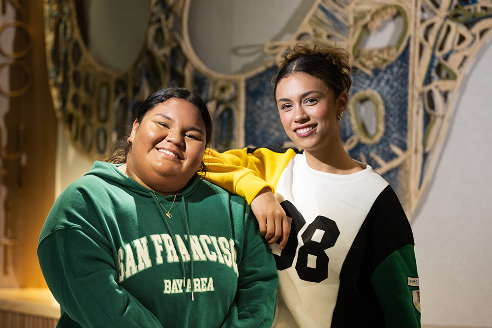 Two Indigenous Australian young women smile at the camera in front of a woven artwork. One woman wears a green sweatshirt while the other wears a yellow, black and green football jersey.
