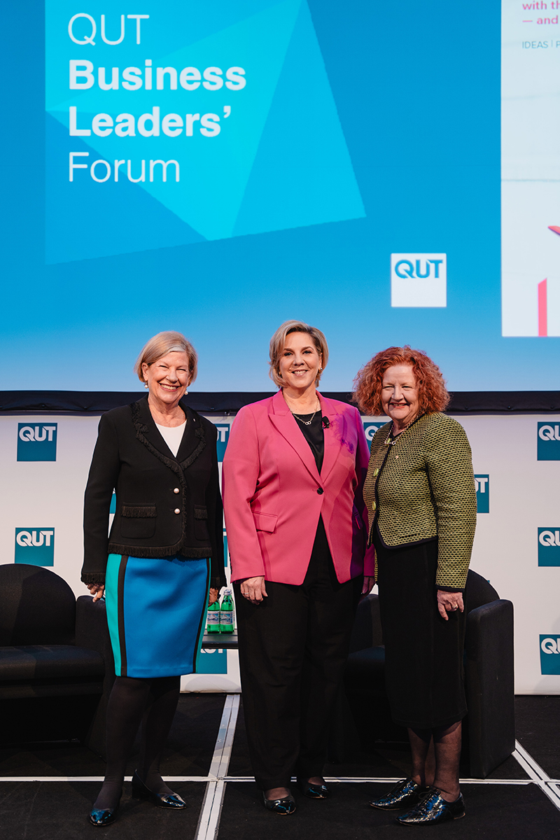 Three women stand on stage and smile at the camera. Behind them a sign reads QUT Business Leaders' Forum.